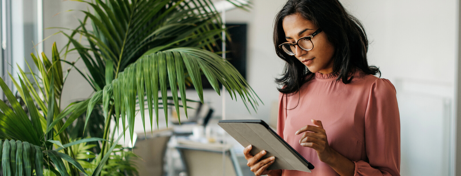 Woman in casual business attire using a tablet device near a plant